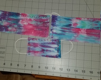 May include: Three face masks with tie-dye patterns in shades of pink, blue, and purple. The masks are made of fabric and have white elastic ear loops. The masks are laid out on a gray surface with a ruler.