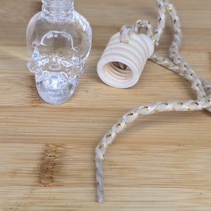 May include: A clear glass skull-shaped bottle with a wooden cap and a white cord with gold accents. The bottle is a car air freshener.