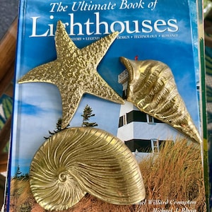 Può includere: Tre oggetti decorativi color ottone a forma di stella marina, conchiglia a spirale e conchiglia su un libro intitolato The Ultimate Book of Lighthouses. Gli oggetti in metallo presentano superfici testurizzate e una finitura dorata.