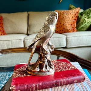 May include: A metallic brass-toned parrot figurine perched on a textured tree stump base. The bird sculpture is positioned on top of a red hardcover book with gold-colored decorative edges, resting on a patterned surface.