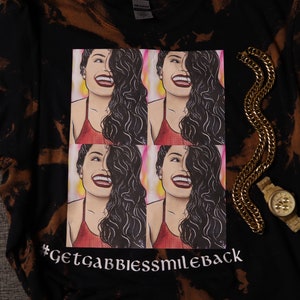 May include: Black tie-dye t-shirt with a white graphic of four images of a woman with long dark hair smiling. The text "#GetGabbiessmileback" is printed in white at the bottom of the graphic.