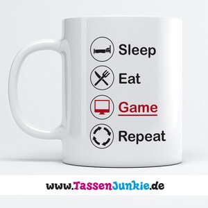 Könnte beinhalten: Weißer Keramikbecher mit gebogenem Henkel. Der Becher zeigt die Worte "Sleep, Eat, Game, Repeat" in schwarzer Schrift mit einem roten "Game" und entsprechenden Symbolen. Die Website-Adresse "www.TassenJunkie.de" ist unten aufgedruckt.