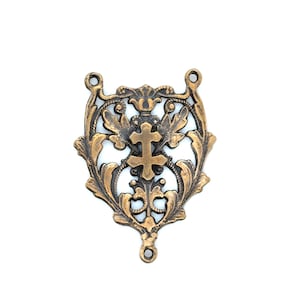 May include: Ornate bronze-colored metal pendant with a central cross design. The pendant features intricate floral and scrollwork patterns, with three small loops for attachment. The overall design is symmetrical and detailed, suitable for jewelry making or decorative purposes.