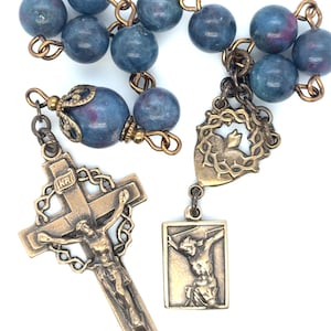 May include: A rosary featuring round blue and magenta stone beads connected by bronze-toned metal links. It includes a bronze crucifix with the letters INRI at the top, a shield-shaped medal depicting a heart with thorns, and a rectangular medal.