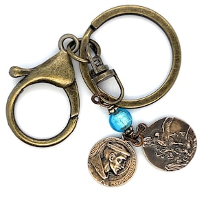 May include: Antique bronze-toned keychain with a lobster clasp and a large key ring. It features a blue bead and two circular charms. One charm depicts a religious figure, and the other shows an angel. The keychain is designed with a vintage aesthetic.