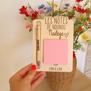 May include: A wooden notepad holder with a pink sticky note, a pen, and the text "LES NOTES DE NOUNOU Nad&egrave;ge". The pen is engraved with "NOUNOU NAD&Egrave;GE". The notepad holder is held in front of a vase of flowers.