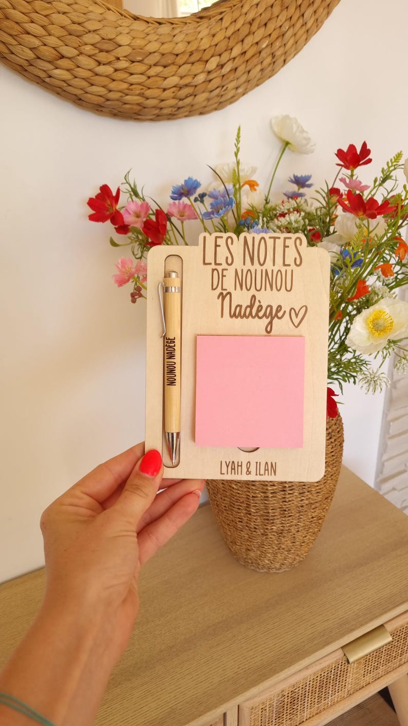 May include: A wooden notepad holder with a pink sticky note, a pen, and the text "LES NOTES DE NOUNOU Nad&egrave;ge". The pen is engraved with "NOUNOU NAD&Egrave;GE". The notepad holder is held in front of a vase of flowers.
