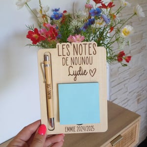 May include: Wooden memo board with a pen and a blue sticky note. The board has the text "LES NOTES DE NOUNOU Lydie ♡" and "EMMIE 2024/2025". A pen with the text "NOUNOU Lydie" is in the holder. A vase of colorful flowers is in the background.
