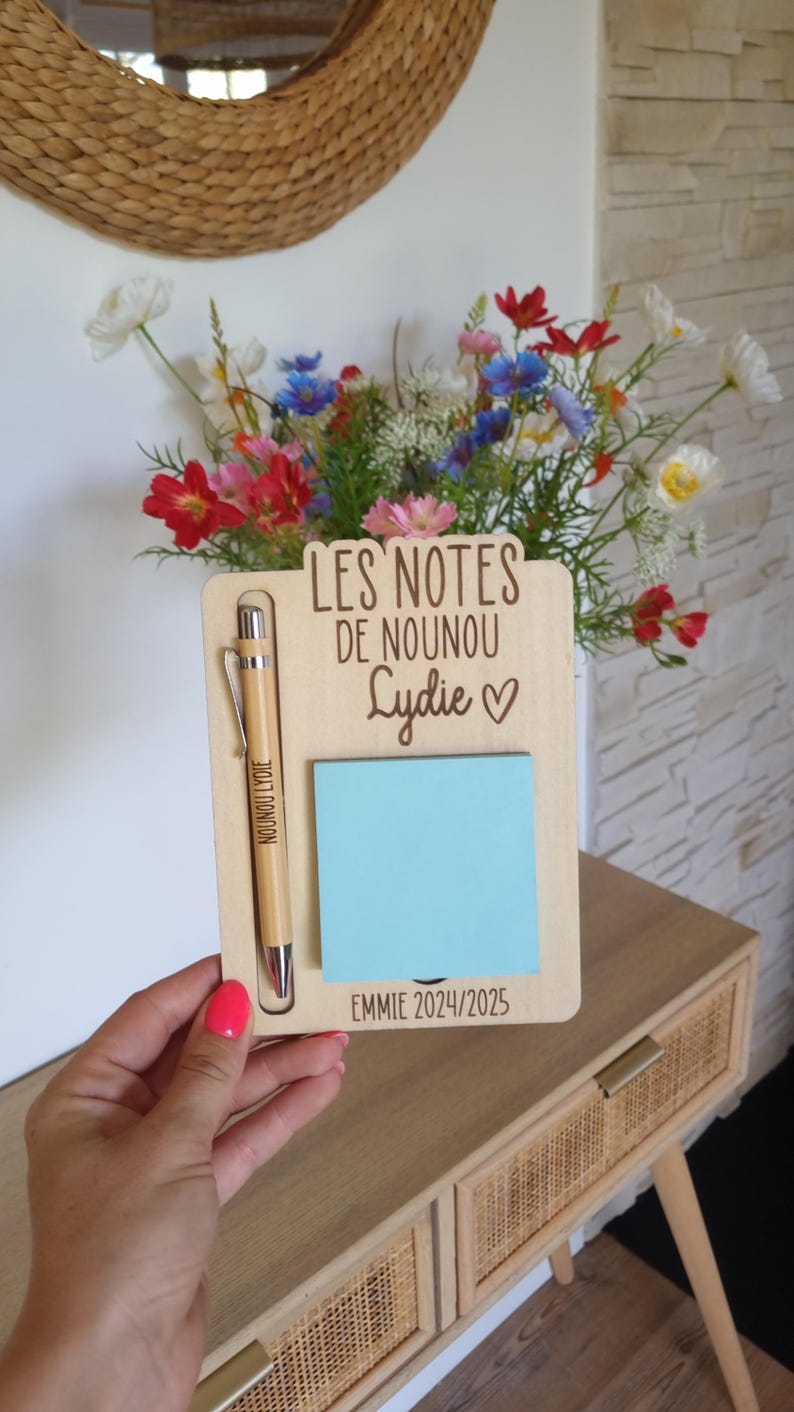 May include: Wooden memo board with a pen and a blue sticky note. The board has the text "LES NOTES DE NOUNOU Lydie ♡" and "EMMIE 2024/2025". A pen with the text "NOUNOU Lydie" is in the holder. A vase of colorful flowers is in the background.