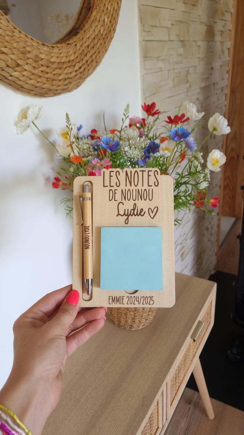Peut inclure: Un tableau mémo en bois avec un post-it bleu clair, un stylo en bois et le texte "LES NOTES DE NOUNOU Lydia ♡" et "EMMIE 2024/2025". Un bouquet de fleurs colorées est en arrière-plan.