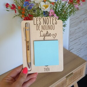 May include: A wooden notepad holder with a pen and a blue sticky note. The notepad holder has the text "LES NOTES DE NOUNOU Lydie" and the name "Th&eacute;a" at the bottom. A bamboo pen is in the holder. A bouquet of colorful flowers is in the background.