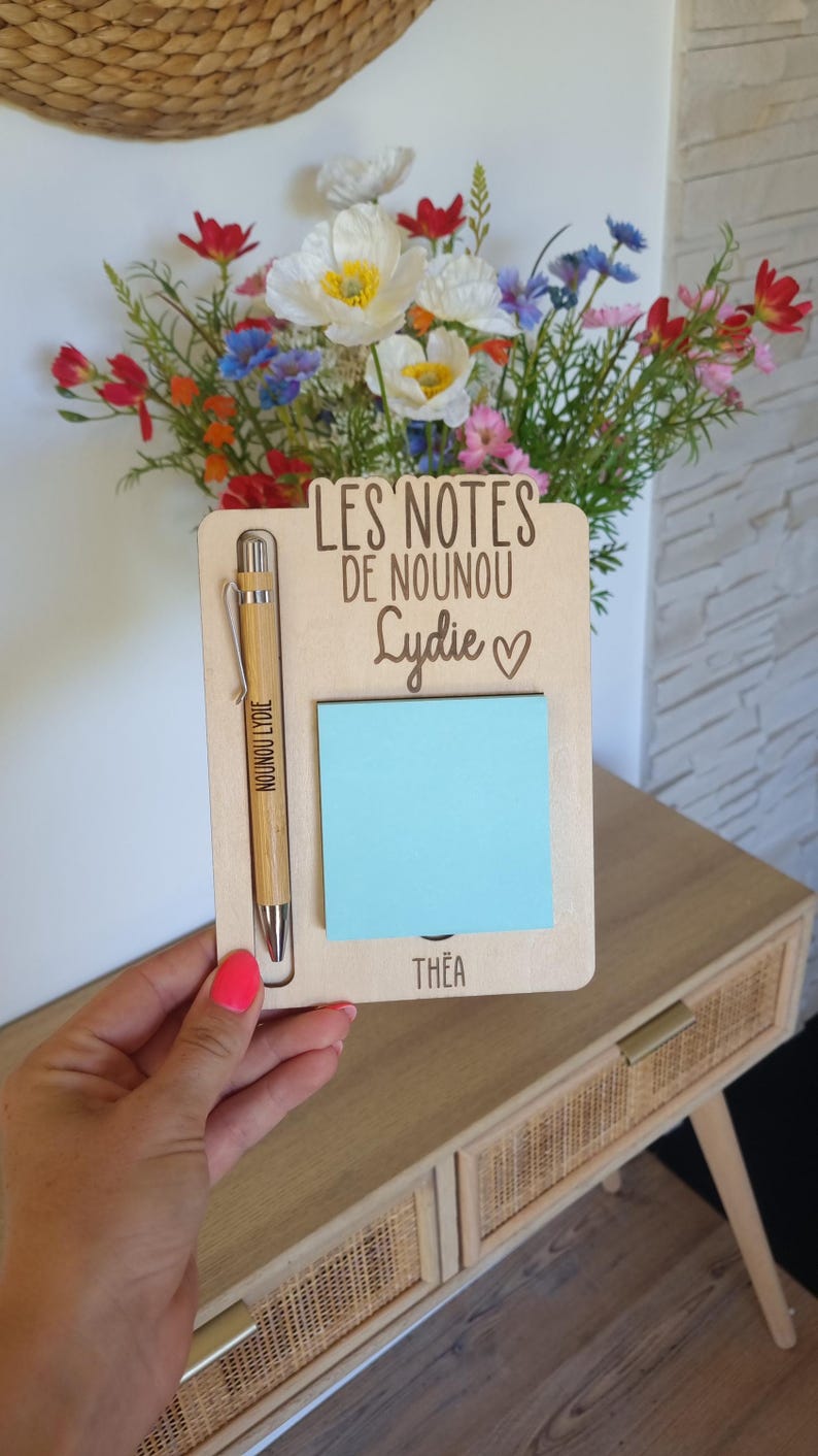 May include: A wooden notepad holder with a pen and a blue sticky note. The notepad holder has the text "LES NOTES DE NOUNOU Lydie" and the name "Th&eacute;a" at the bottom. A bamboo pen is in the holder. A bouquet of colorful flowers is in the background.