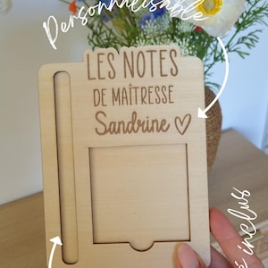 May include: A light-colored wooden notepad holder with the text "LES NOTES DE MA&Icirc;TRESSE Sandrine ♡". The holder has a slot for a pen and a rectangular space for notes. A bouquet of colorful flowers is in the background.