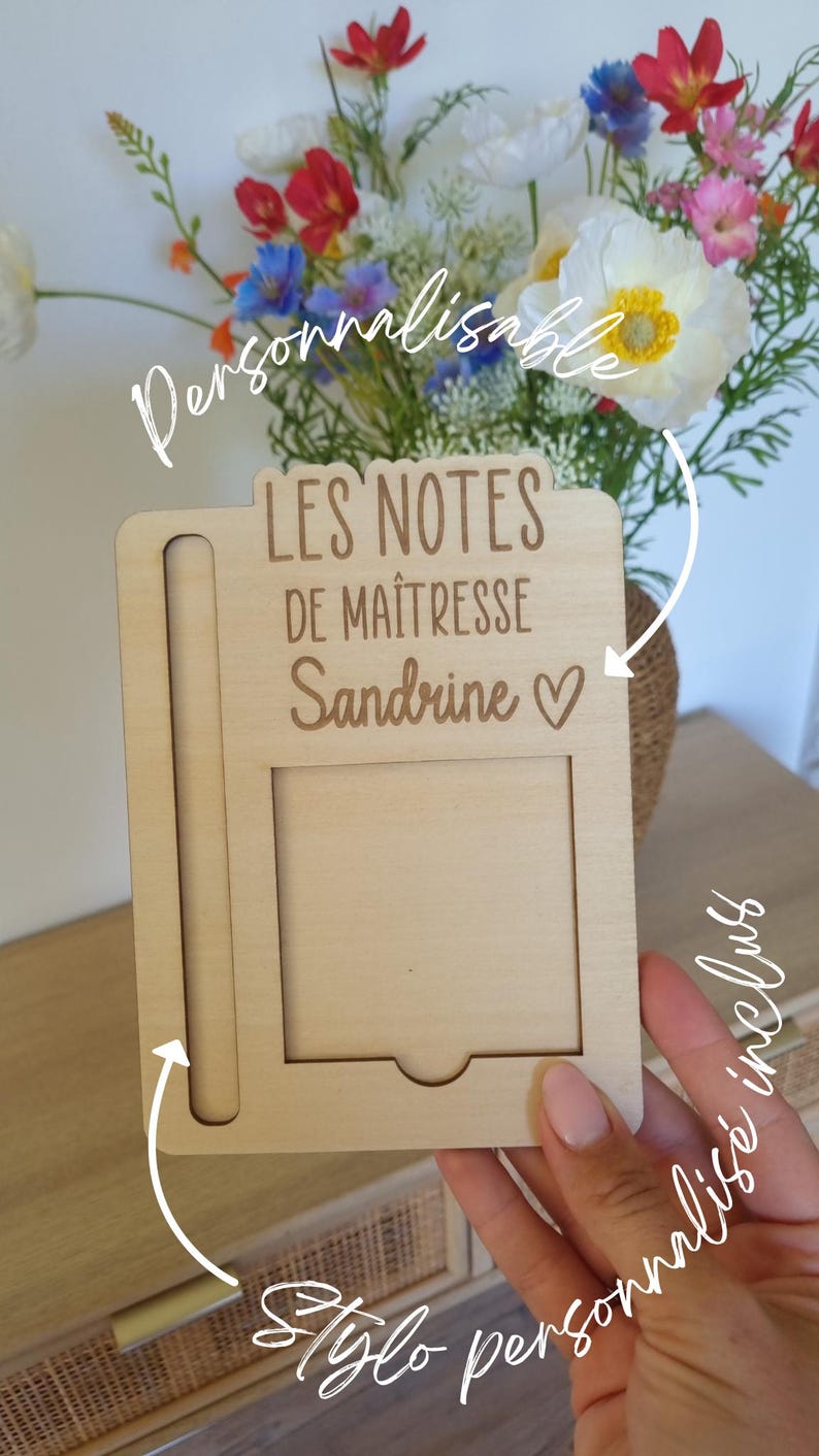 May include: A light-colored wooden notepad holder with the text "LES NOTES DE MA&Icirc;TRESSE Sandrine ♡". The holder has a slot for a pen and a rectangular space for notes. A bouquet of colorful flowers is in the background.