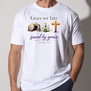 May include: White t-shirt featuring the text "Grace not fate" above an illustration of a tomb, flowers, and a cross. The words "saved by grace" are written in a cursive font, with "Romans 11:6" below.