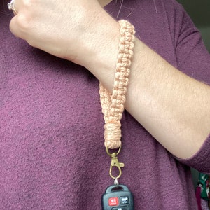 May include: A peach-colored braided lanyard with a gold key chain and a black car key with buttons. The lanyard is worn around a person's wrist.