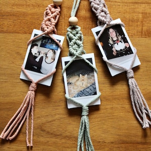 May include: Three macrame photo holders with wooden beads and tassels. Each holder features a different photograph, including a couple, a cat, and a woman holding a cake.