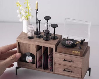 Può includere: Mobile giradischi in miniatura in legno con un piccolo giradischi, cassetti e un vano per dischi in vinile. Il mobile è decorato con piccoli candelabri neri, un vaso di fiori bianchi e una campana di vetro trasparente. Il giradischi ha il testo "IN BOX STUDIO".