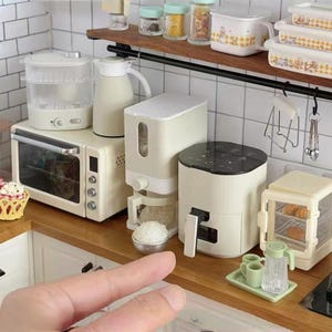 May include: A miniature kitchen set with a white oven, a white coffee maker, a white air fryer, a white rice cooker, and a white steamer. The appliances are arranged on a wooden countertop with a white tiled backsplash.