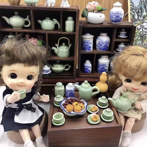 May include: A miniature tea set with a teapot, cups, and saucers, along with a plate of mooncakes. The set is arranged on a wooden table with two dolls in the background.