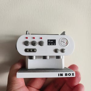 May include: White miniature espresso machine with a silver trim and a digital display showing "TIME 13:09". The machine has a silver gauge and three silver spouts. The base of the machine is white with the text "IN BOX" printed in black.