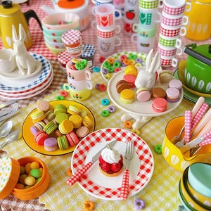 May include: A miniature tea party set with a checkered tablecloth, colorful teacups, plates, and macarons. There are also miniature cakes, a teapot, and a pitcher.