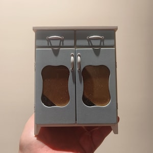 May include: A small, light blue cabinet with two drawers and two doors. The doors have white handles and glass panels with a scalloped edge. The cabinet is sitting on a hand.