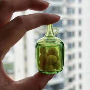 Puede incluir: Una pequeña botella de vidrio verde con un tapón de corcho y un asa de metal. La botella está llena de pequeños caramelos amarillos con forma de limón.