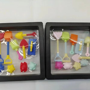 May include: Two black frames with clear plastic inserts, each containing a set of colourful plastic beach toys. The left frame has a red, orange, yellow, green, blue, and pink set. The right frame has a pink, green, orange, blue, and yellow set.