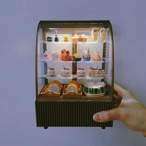 May include: A miniature glass display case filled with various miniature cakes, cupcakes, and pastries. The case has three shelves and a brown wooden base. The case is lit from the inside.