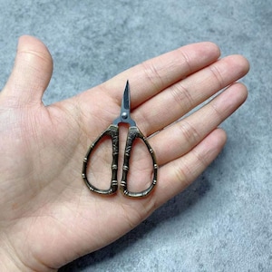 May include: A pair of small, antique-style scissors with a gold finish and a bamboo design. The scissors have a black blade and are held in a hand.
