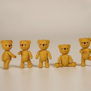 Puede incluir: Cinco pequeños osos de peluche amarillos con ojos y narices marrones. Los osos están de pie en fila, con el primer oso mirando hacia la izquierda, el segundo oso mirando hacia la derecha, el tercer oso mirando hacia adelante, el cuarto oso sentado con las piernas abiertas y el quinto oso mirando hacia la derecha con una pierna levantada.