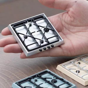 May include: Three miniature stovetops in different colors: silver, light blue, and cream. Each stovetop has four black burner grates.