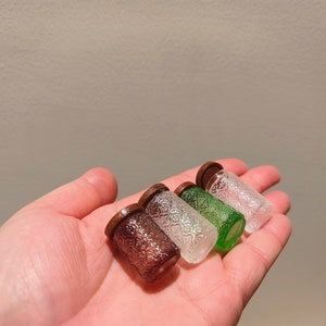 Op de afbeelding: Vier kleine glazen potjes met houten deksels. De potjes zijn helder, groen en bruin. De potjes zijn in een rij op een hand gerangschikt.