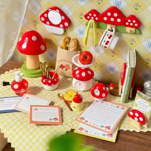 May include: A miniature dollhouse scene with a red and white mushroom theme. The scene includes a mushroom lamp, a mushroom scale, a mushroom hook, and a mushroom notepad holder. There are also miniature food items, such as bread and an apple.