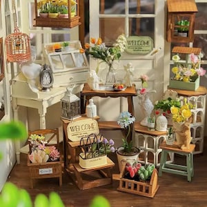 Peut inclure: Une maison de poupée miniature sur le thème d'une boutique de fleurs. La maison de poupée comprend une variété de meubles miniatures, de plantes et d'accessoires, notamment une pancarte de bienvenue, une petite table avec un vase de fleurs, une cage à oiseaux et une serre miniature. La maison de poupée est peinte dans une couleur bois clair et a une porte blanche avec une fenêtre.