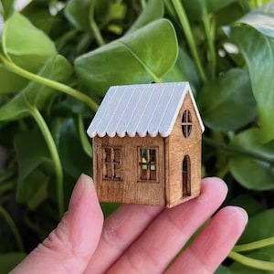 May include: A miniature wooden house with a white roof and a brown wooden door. The house has two windows with white frames. The house is sitting on a person's hand.