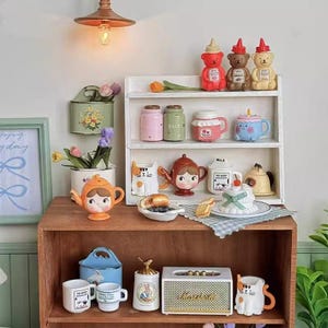 以下が含まれることがあります： 木製の棚には、様々な装飾品が飾られています。クマの形をした調味料ボトル、ティーポット、マグカップ、ジャーがあります。オレンジ、ピンク、ブルーなど、様々な色があります。小さなケーキと食べ物のプレートも表示されています。