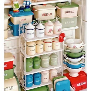 May include: A white metal shelf unit with various miniature kitchen items, including bread boxes, jars labeled "sugar", "salt", and "tea", and bowls in various colors. The shelf unit is on a white countertop with a red and white checkered tablecloth.