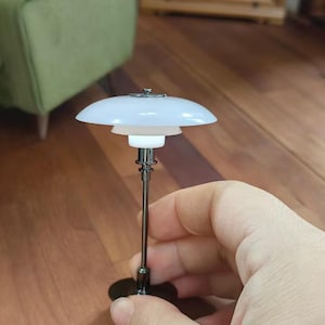 以下が含まれることがあります： 丸い平らなシェードと黒い金属製のベースが付いた白いテーブルランプ。ランプは木製の表面の上に置かれています