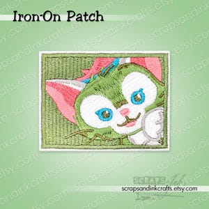 May include: Embroidered iron-on patch featuring a cartoon cat with green fur, pink ears, and blue eyes. The patch is rectangular with a green background and white border. Text "Iron-On Patch" is at the top.