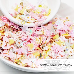 May include: A bowl filled with pink, yellow, and white glitter and resin shapes. The shapes include flowers, butterflies, stars, and hearts. The glitter is iridescent and catches the light. The bowl is white and has a lip around the edge. The text "SCRAPS & INK" is visible on the bottom of the bowl.