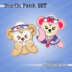 May include: An iron-on patch set featuring two embroidered bear characters. One tan bear wears a white sailor hat with a red heart icon. The other pink bear wears a white sailor hat with a red bow and a dark blue sailor collar with a red tie.