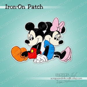Può includere: Toppa termoadesiva ricamata con Topolino e Minnie. Topolino indossa pantaloncini rossi e scarpe gialle, Minnie un vestito blu e scarpe rosa. La toppa ha un bordo bianco e il testo "Iron-On Patch".