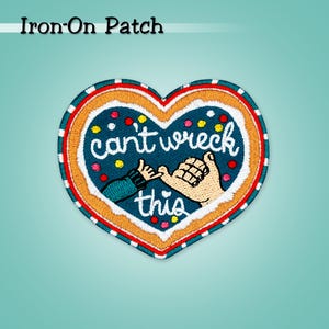 May include: An iron-on patch in a heart shape with a blue background and the words "can't wreck this". The patch features two hands clasped together, with colorful accents. The patch is outlined in red and white.