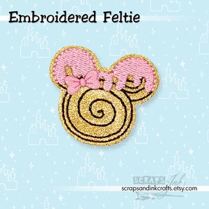 Puede incluir: Fieltro bordado con forma de cabeza de ratón con orejas rosas y un lazo rosa. El centro de la cabeza es purpurina dorada con un diseño en espiral. El texto "Embroidered Feltie" está en la parte superior.