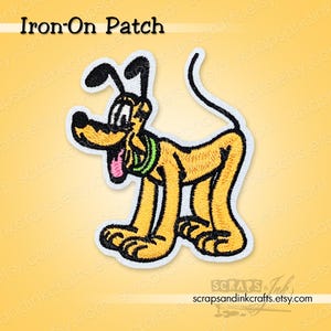Op de afbeelding: Een geborduurde opstrijkbare patch met Pluto, een gele cartoonhond met zwarte oren en een zwarte staart. De patch heeft een witte rand en een groene kraag. De tekst "Iron-On Patch" staat bovenaan.
