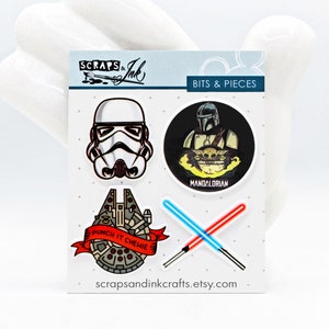 Puede incluir: Un conjunto de cuatro parches termoadhesivos con temática de Star Wars. Los parches presentan un casco de Stormtrooper, un Mandaloriano con Grogu, un Halcón Milenario con el texto "Punch it Chewie" y dos sables de luz cruzados. Los parches están sobre un fondo blanco con el texto "Scraps & Ink" y "Bits & Pieces" en la parte superior.