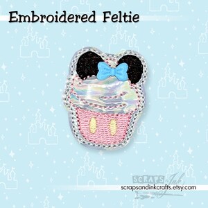 May include: Embroidered feltie in the shape of a cupcake with Mickey Mouse ears and a blue bow. The cupcake has a pink bottom with two yellow ovals, a pearlescent top, and black ears. The feltie is outlined with silver stitching.