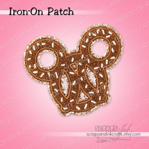 May include: An iron-on patch in the shape of a pretzel with Mickey Mouse ears. The patch is brown with white sprinkles and a gold glitter border. The text "Iron-On Patch" is at the top.